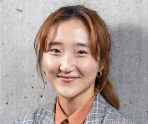 An Asian woman with light brown hair tied back wears gold hoops and a grey plaid blazer against a concrete wall.