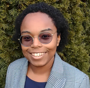 A Black woman with short natural hair, wearing round tinted glasses and a grey plaid blazer in front of a green hedge.