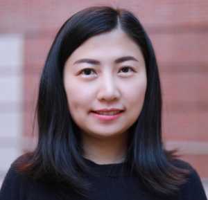 An Asian woman with long dark hair in a black top, standing in front of a blurred red brick wall.