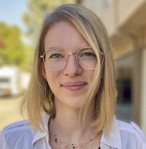 A White woman with shoulder-length blonde hair, gold hexagonal glasses, and a white shirt, standing outdoors.