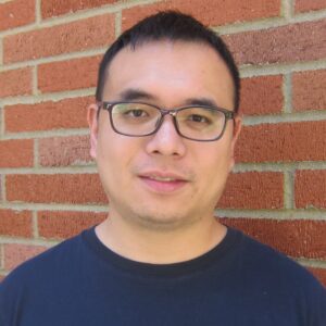 An Asian man with short hair and rectangular glasses wears a navy t-shirt, standing in front of a red brick wall.