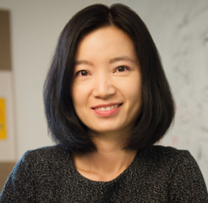 An Asian woman with shoulder-length dark hair wears a textured black top, standing in front of a whiteboard.