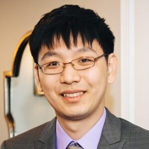 An Asian man with short dark hair and rectangular glasses, wearing a grey suit and lavender shirt indoors.
