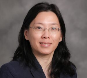 An Asian woman with long dark wavy hair and thin wire-rimmed glasses, wearing a navy blazer against a grey background.
